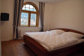 A bedroom with a wooden bed, a window with curtains, a radiator, and a wall-mounted television. - Vacation Rental: 65 square meters, separate bedroom and balcony, Apt. 7 (Aschau im Chiemgau)