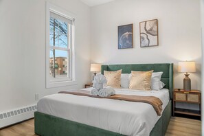 A neatly made bed with a green headboard, two bedside lamps, and a window with a view of buildings. - Edmonton Convention Centre Studio | Parking, Wifi (Edmonton)