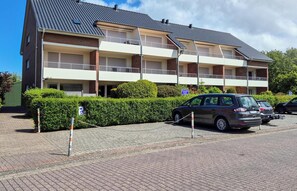 Ein mehrgeschossiges Wohnhaus mit Ziegeldach, Balkonen und einem Parkplatz mit Autos.