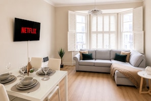 A modern living room with a flat-screen TV mounted on the wall, a dining area with a table set for two, and a comfortable sofa with a view of the city through large windows with shutters. - Kensington 2BDR flat near Hyde Park & Natural History Museum w/ washer & dryer (London)
