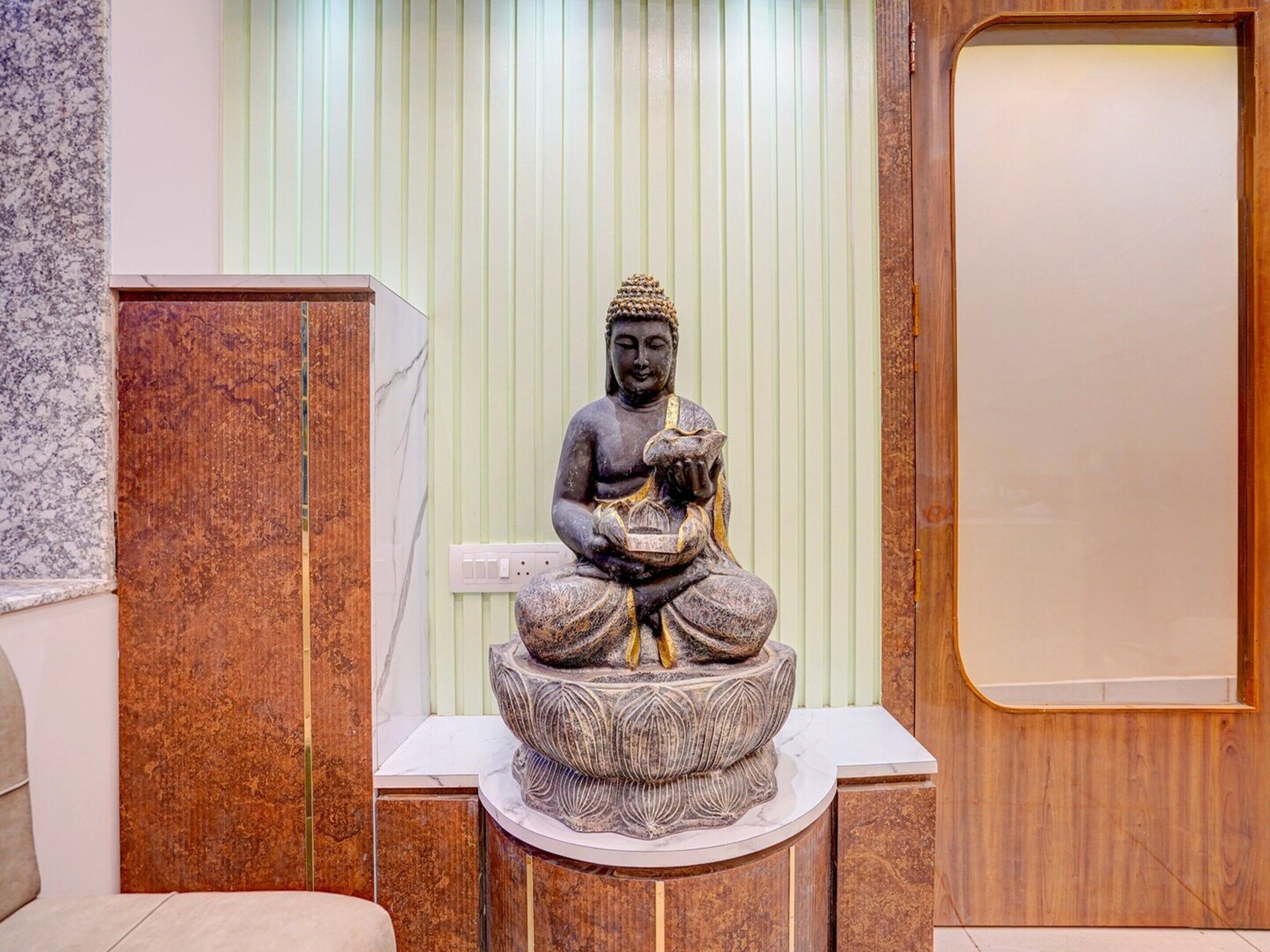 A statue of a seated figure with a bowl, placed on a pedestal in a room with a wooden panel and a marble wall.