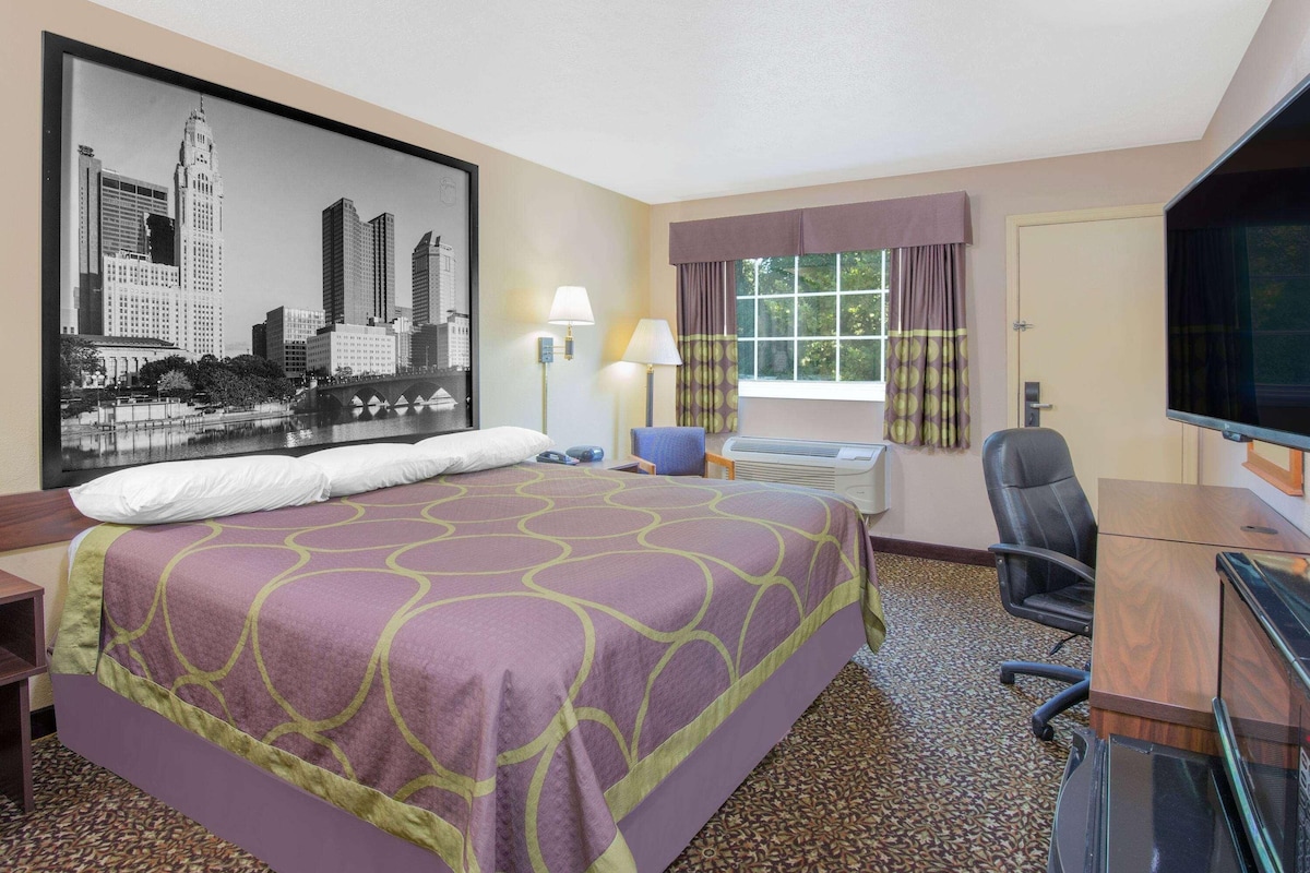Ein Hotelzimmer mit einem großen Bett, einem Schreibtisch mit Stuhl, einem Fernseher und einem Fenster mit Vorhängen.