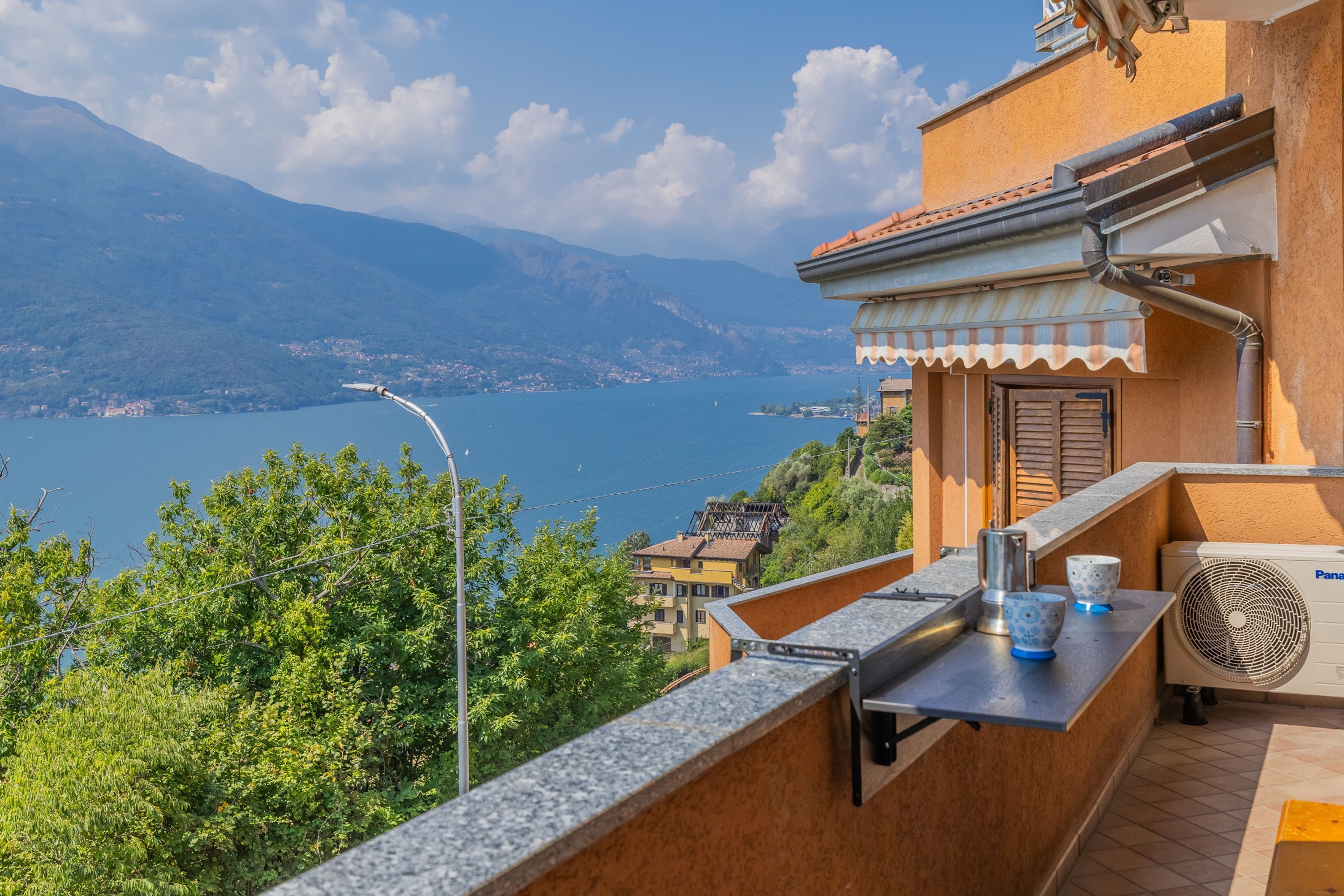 A balcony with a view of a lake and mountains, a table with cups and a kettle, and a window with shutters.