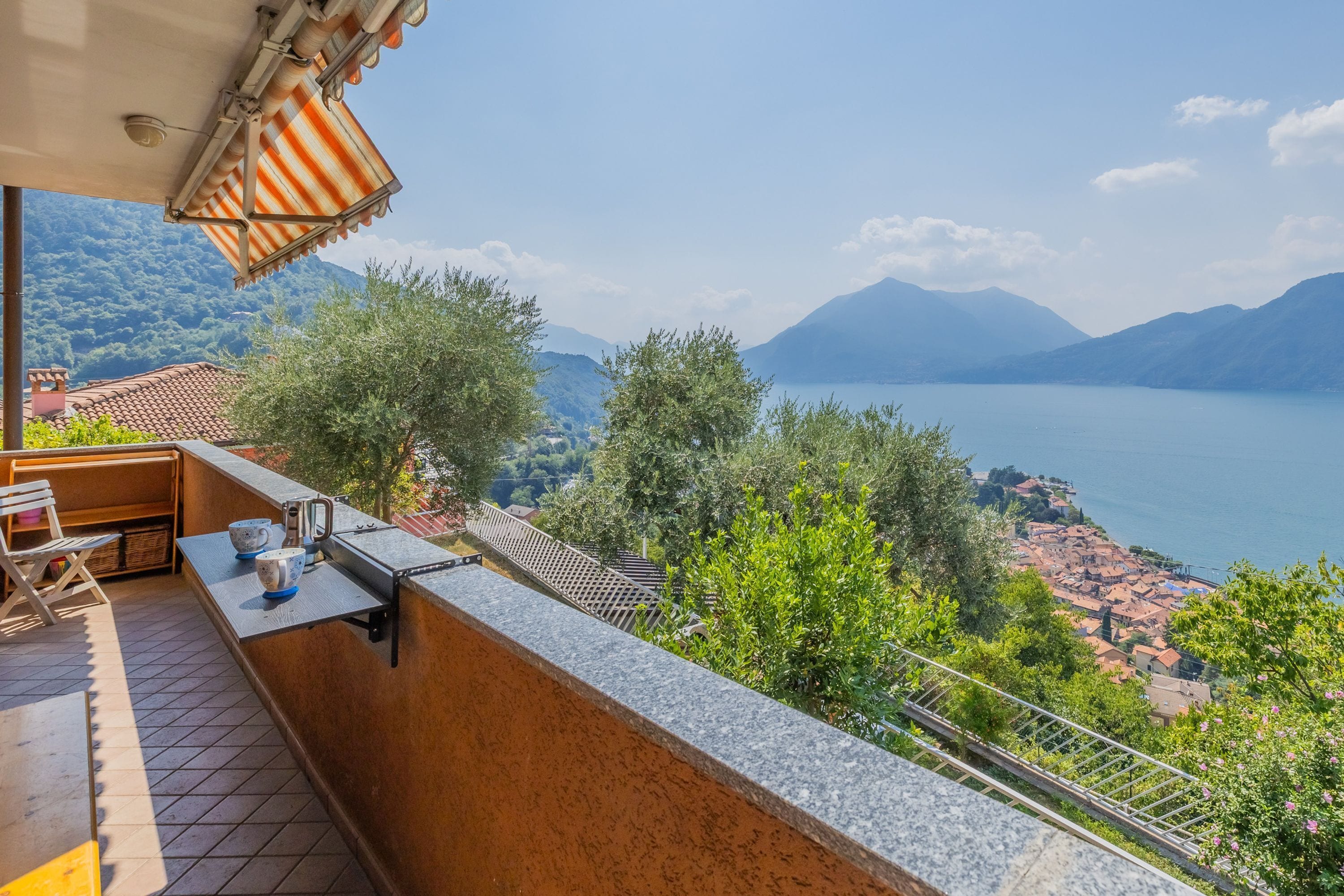 A balcony with a view of a lake and mountains, a table with cups and a kettle, and a chair.