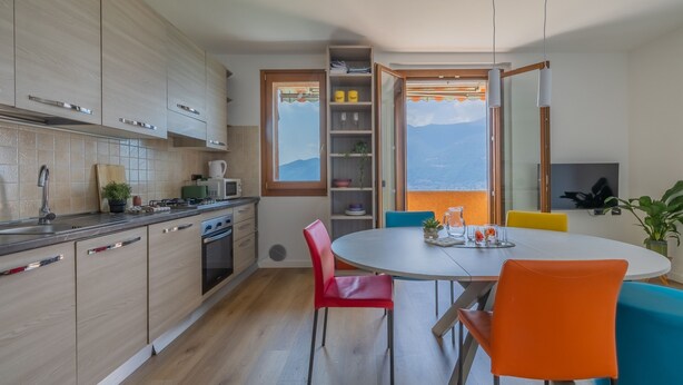 Een moderne keuken met houten kastjes, een eetgedeelte met een ronde tafel en kleurrijke stoelen, en uitzicht op bergen door een raam.