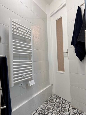 Ein Badezimmer mit weiß gefliester Wand, einer Dusche mit Glastür in Weiß, einem Handtuchhalter mit dunklem Handtuch und einem Bodenbelag mit Muster.