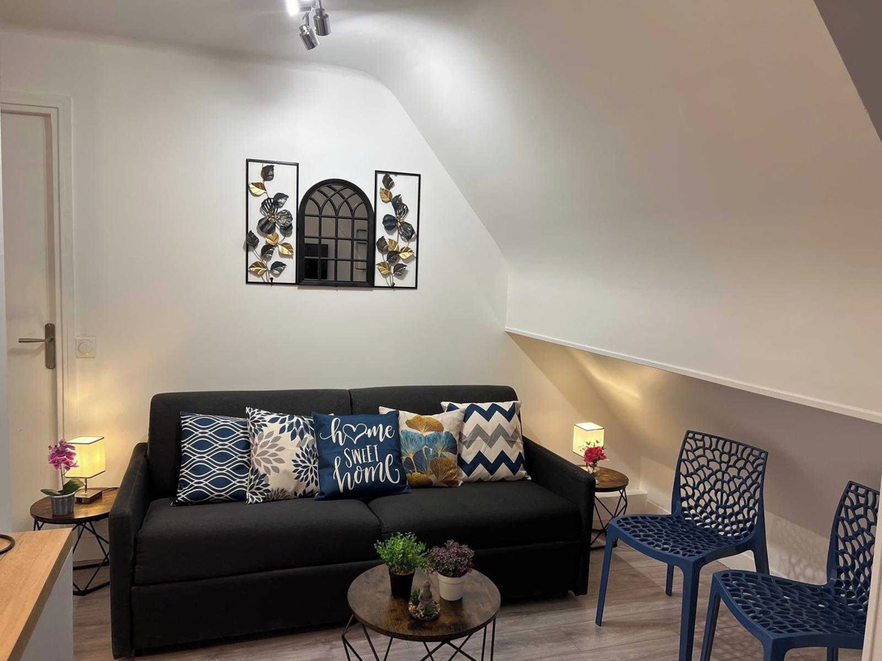 A modern living room with a black sofa, decorative pillows, a small round table with plants, and blue chairs.