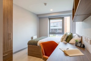 A modern bedroom with a bed, a desk, and a chair. There is a window with a view of buildings outside.
