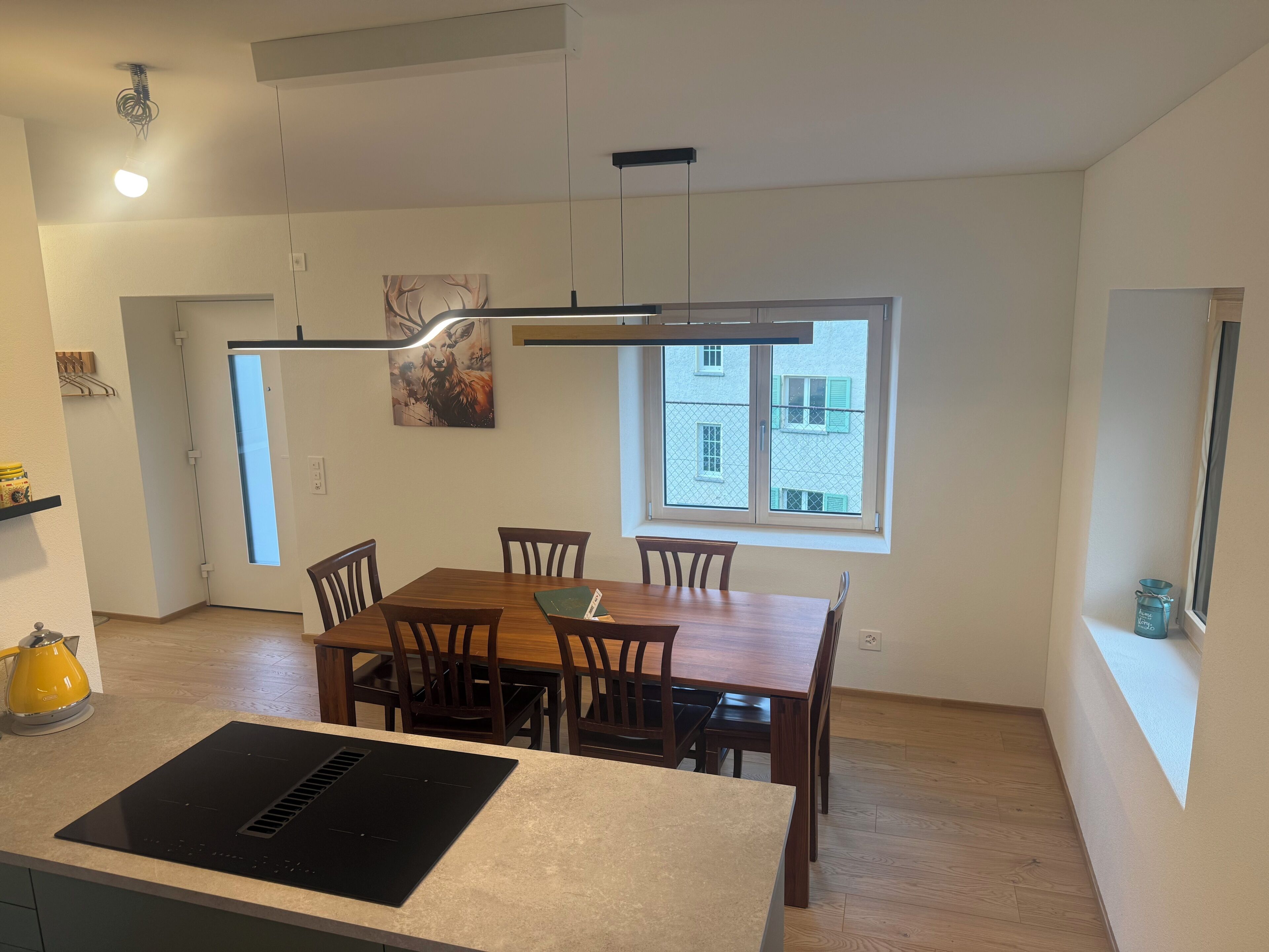 Une cuisine moderne avec un coin repas, des chaises en bois, une plaque de cuisson à induction noire et une fenêtre donnant sur des immeubles.