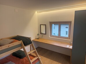 A small, well-lit room with a bunk bed, a desk with a lamp, and a window with a view of buildings.