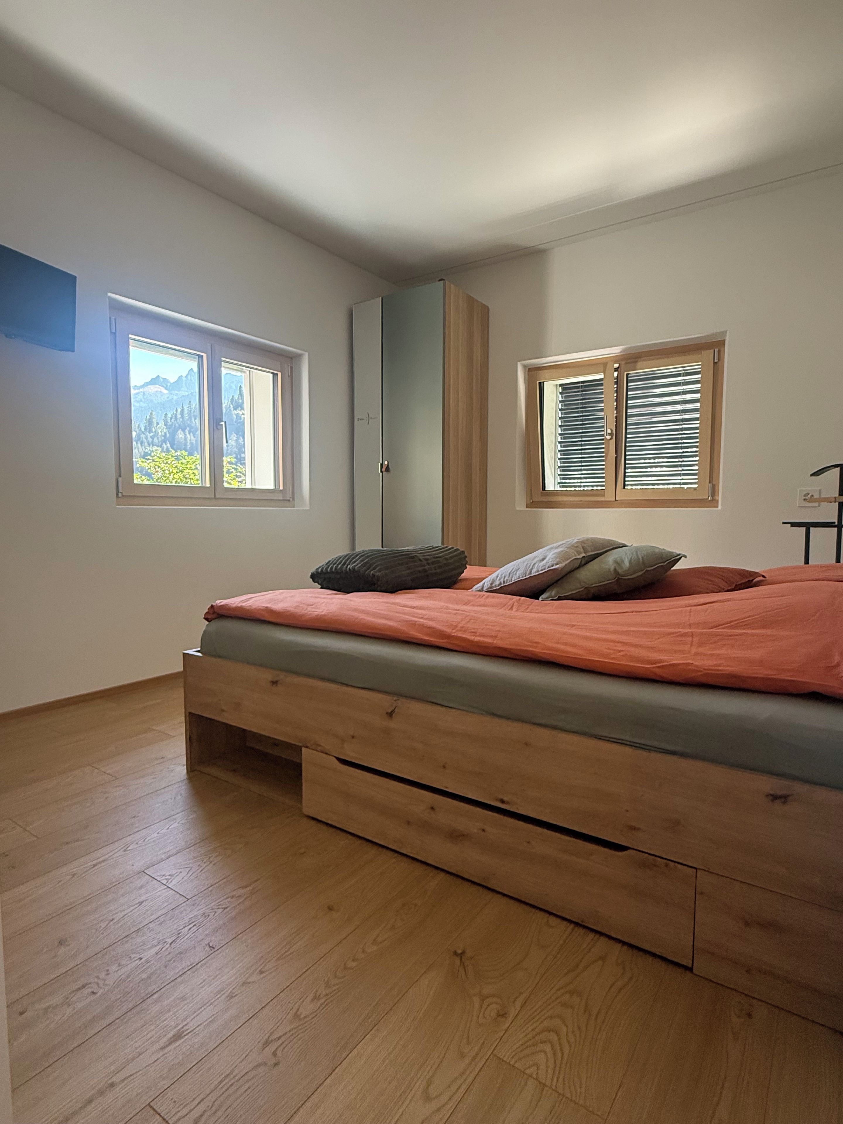 A bedroom with a bed, wooden floor, and two windows with blinds.