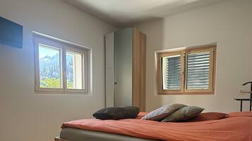A bedroom with a bed, wooden floor, and two windows with blinds.