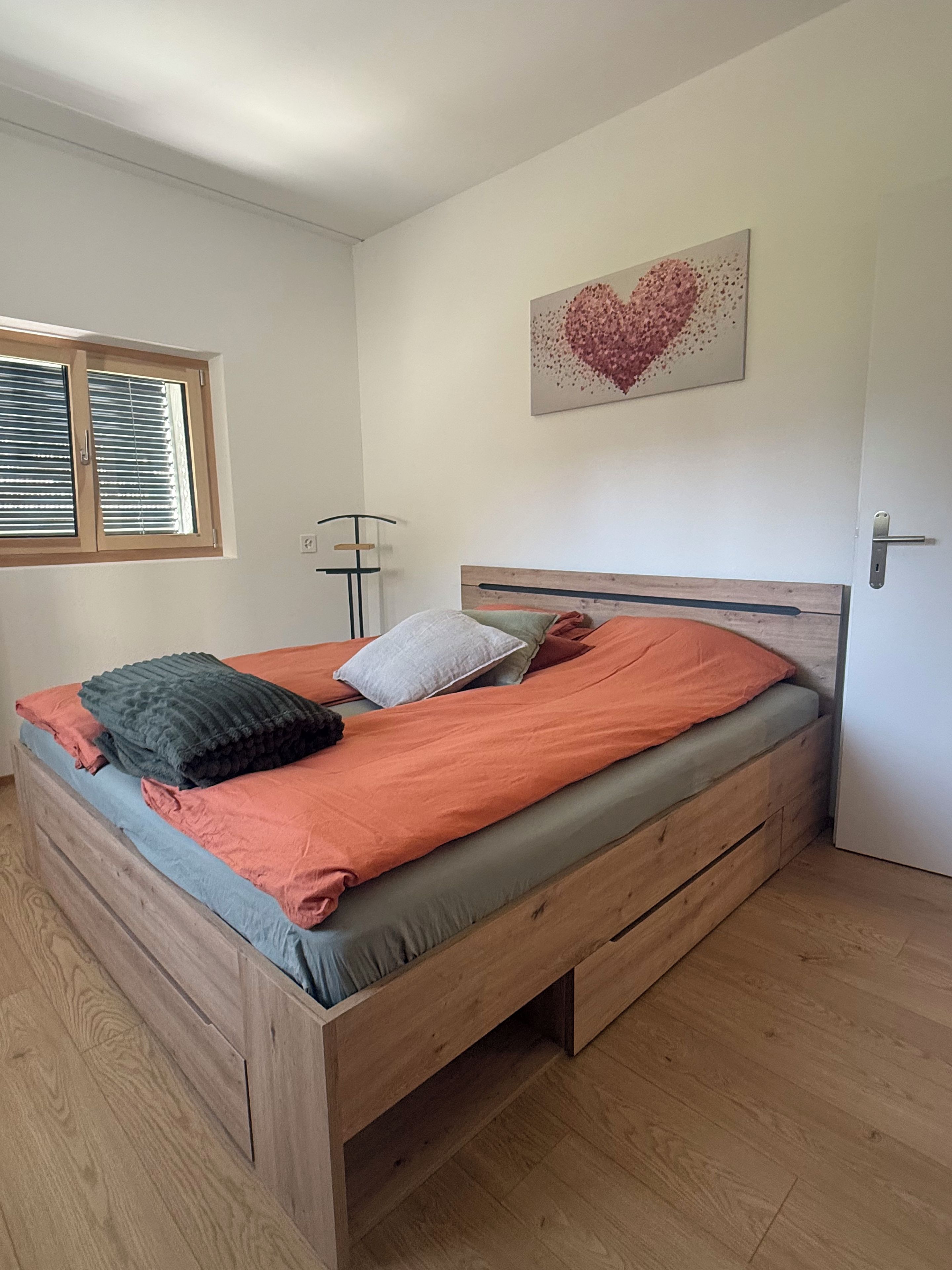 Een slaapkamer met een houten bed, een raam met jaloezieën, een vloerlamp en een hartvormig kunstwerk aan de muur.