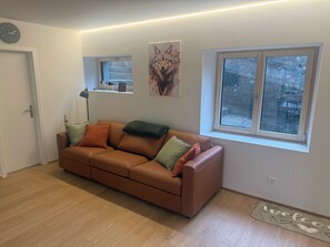 Ein Wohnzimmer mit braunem Ledersofa, einem Fenster mit Blick auf Bäume, einer Wanduhr und einer Fußmatte.