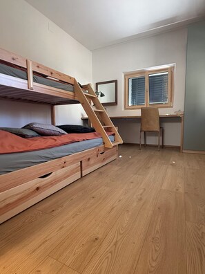Ein Etagenbett mit Holzleiter, ein Schreibtisch mit Stuhl und ein Fenster mit Jalousien.