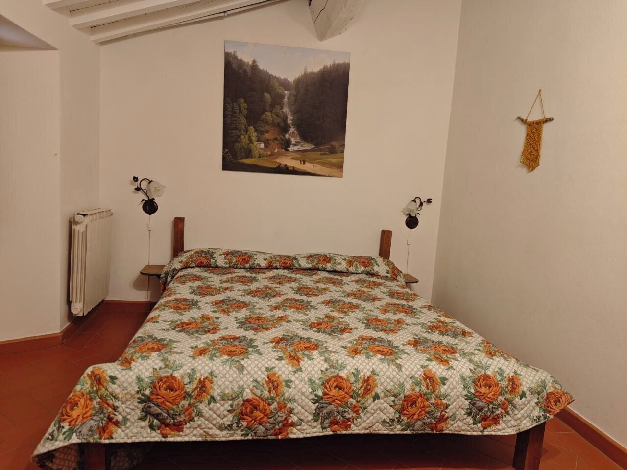 A bedroom with a bed, a radiator, a wall-mounted picture, and a wall-mounted lamp.