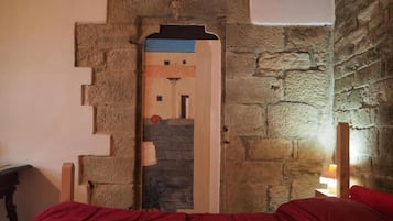 Un dormitorio con pared de piedra, cama con ropa de cama roja y mesita de noche con lámpara.