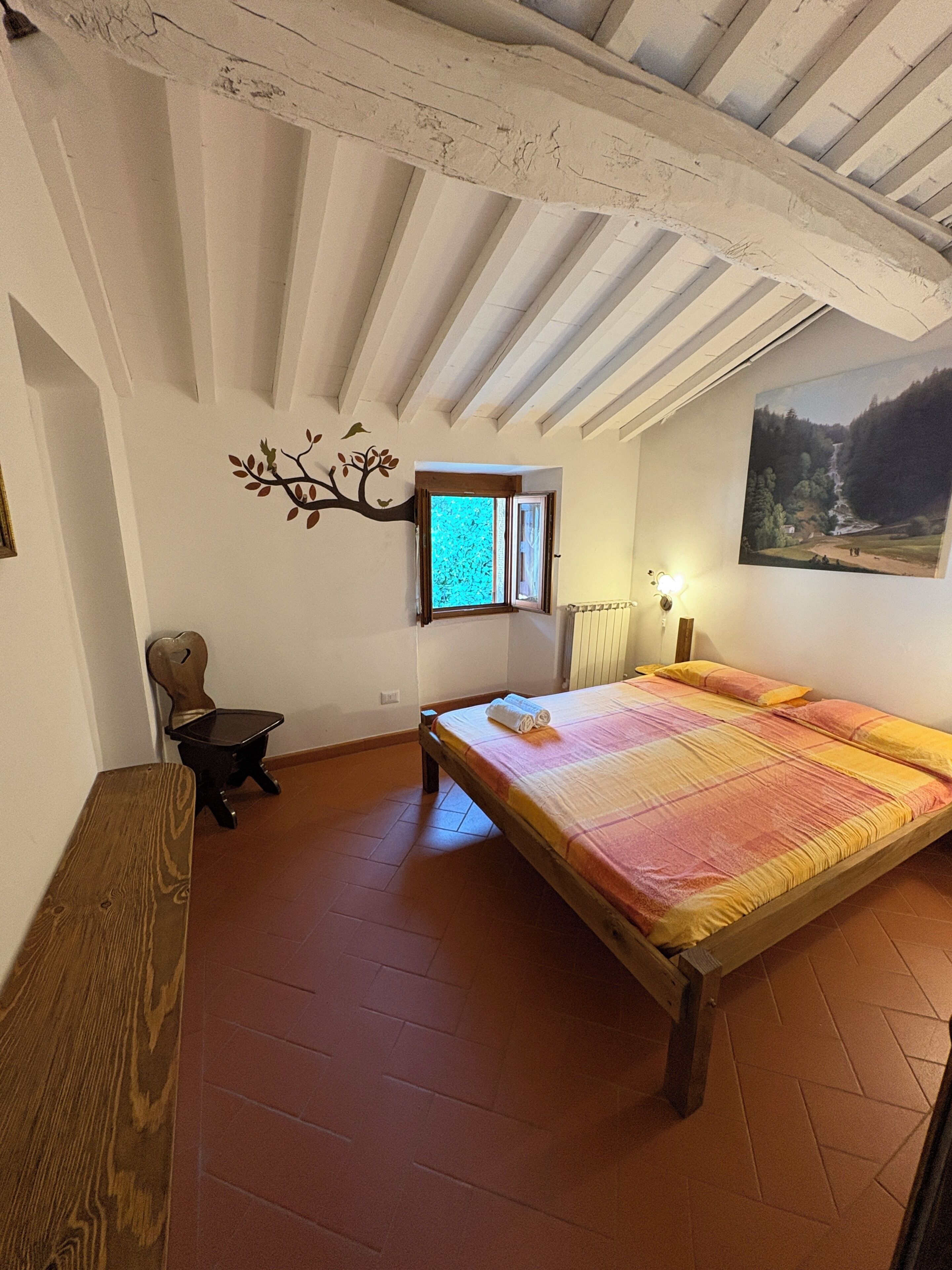 Une chambre à coucher avec un lit en bois, une petite table, une chaise, une fenêtre, un radiateur et un tableau accroché au mur.