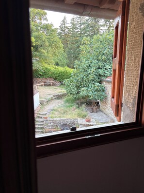 A view through a window with a wooden frame, revealing a garden area with a stone wall, a wooden bench, and various plants. - B&B Antica Foresteria di Tosina (Pelago)