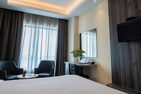 A hotel room with a bed, two chairs, a desk with a laptop, a potted plant, and a TV mounted on the wall.