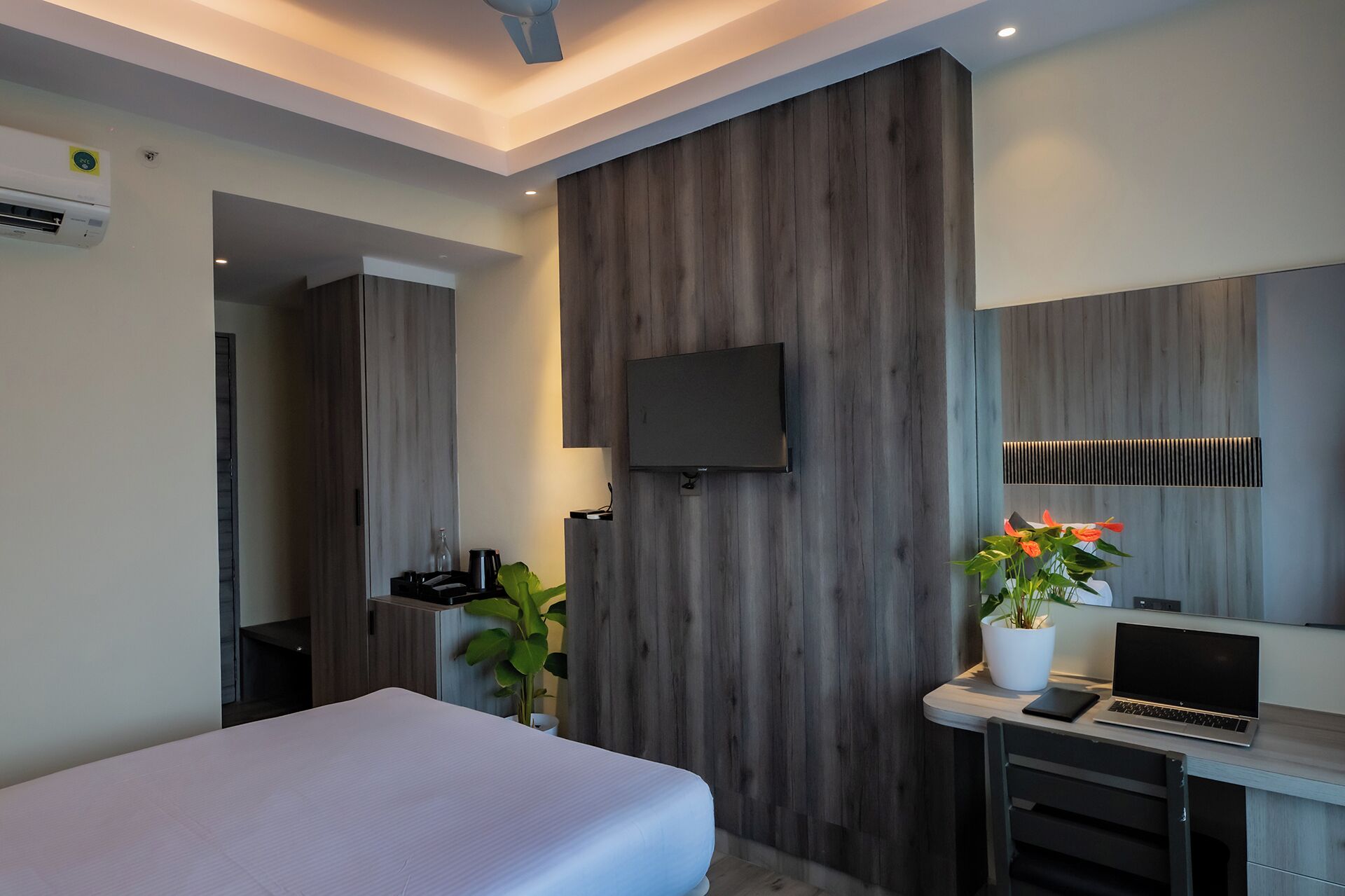 Une chambre d’hôtel moderne équipée d’un lit, d’un bureau avec un ordinateur portable, d’un téléviseur fixé au mur et d’une plante en pot.