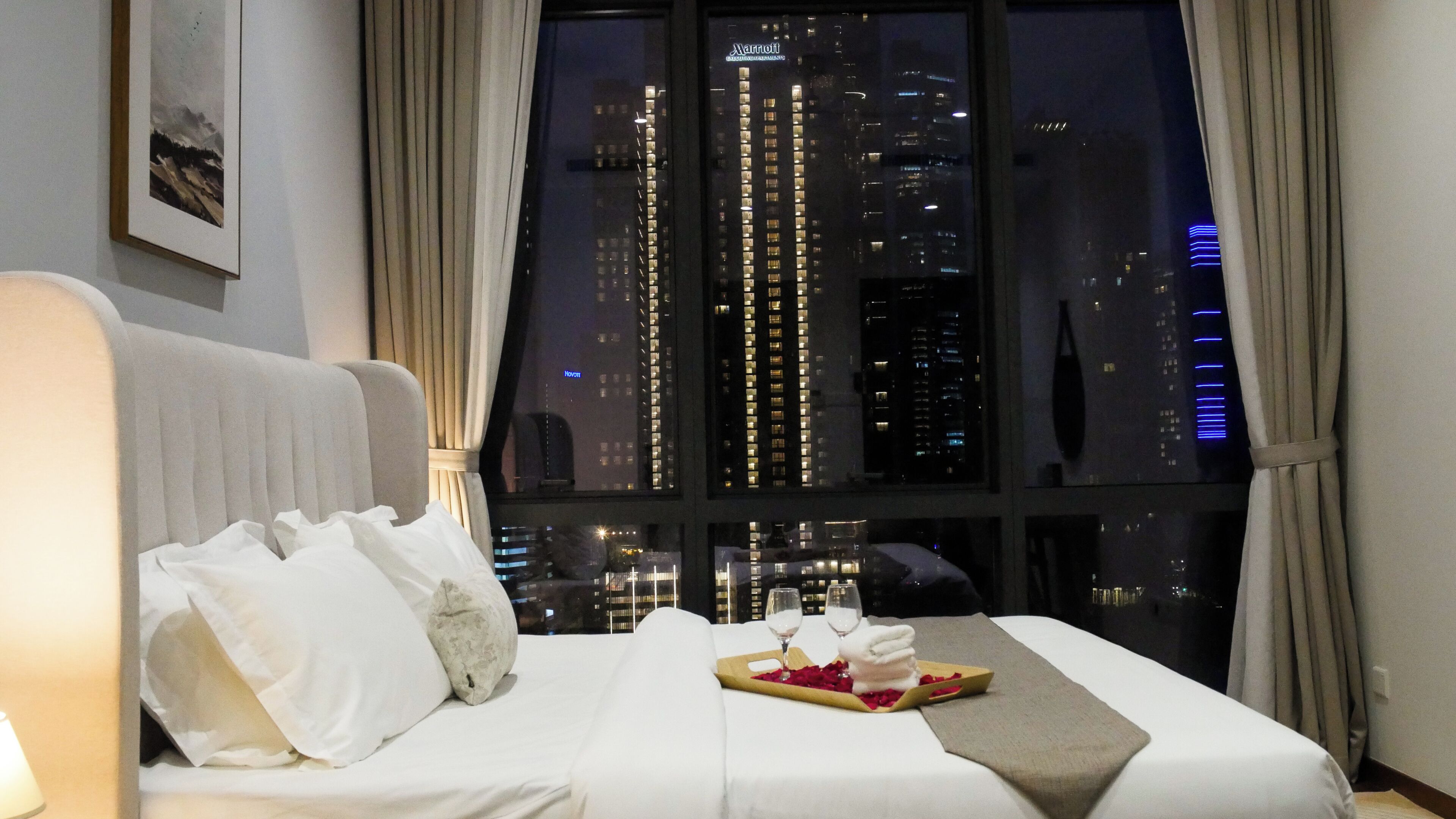 A hotel room with a bed, a window with a view of city buildings, and a framed picture on the wall.