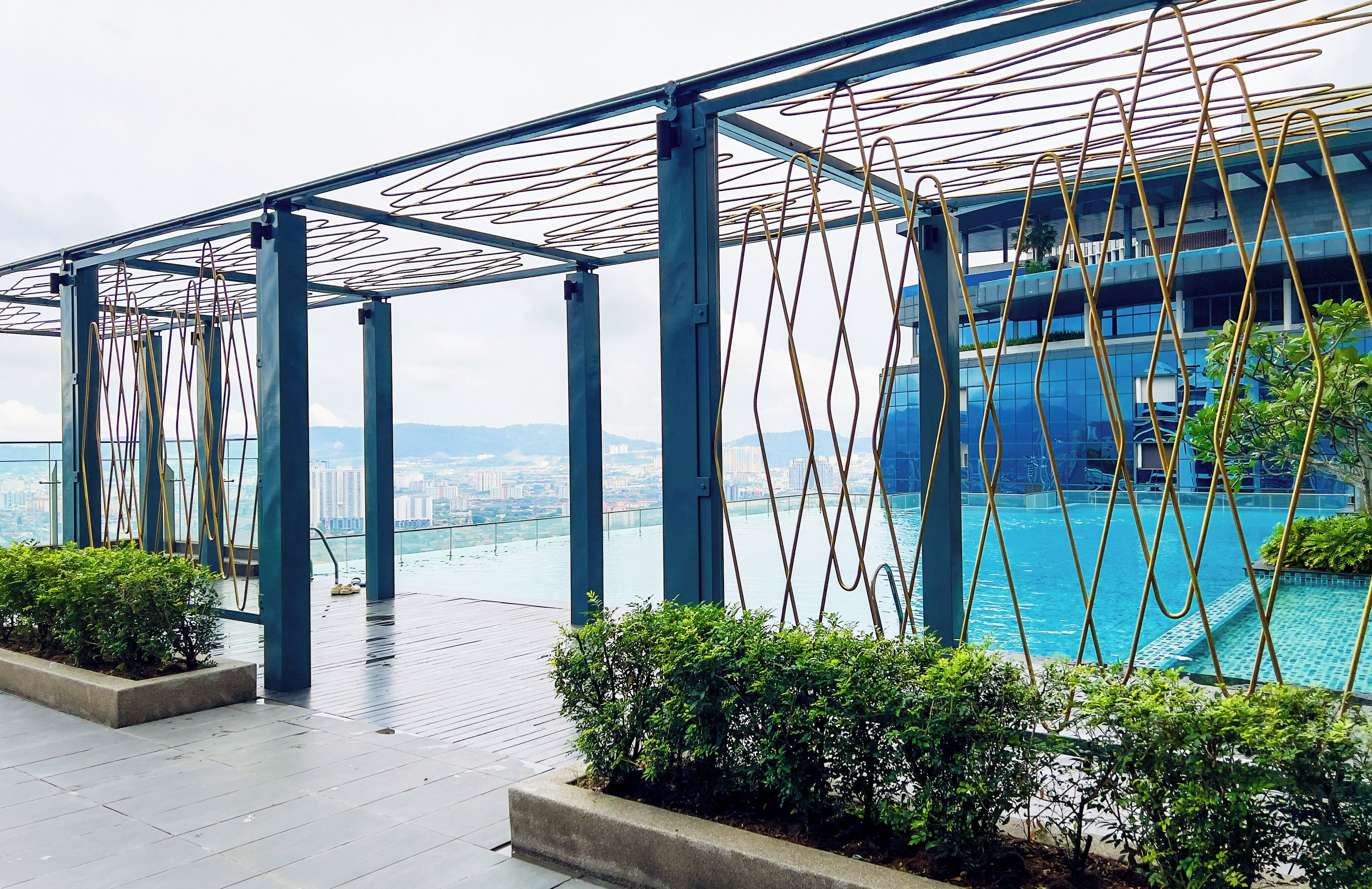 A rooftop pool area with a view of a cityscape, metal railings, and greenery.