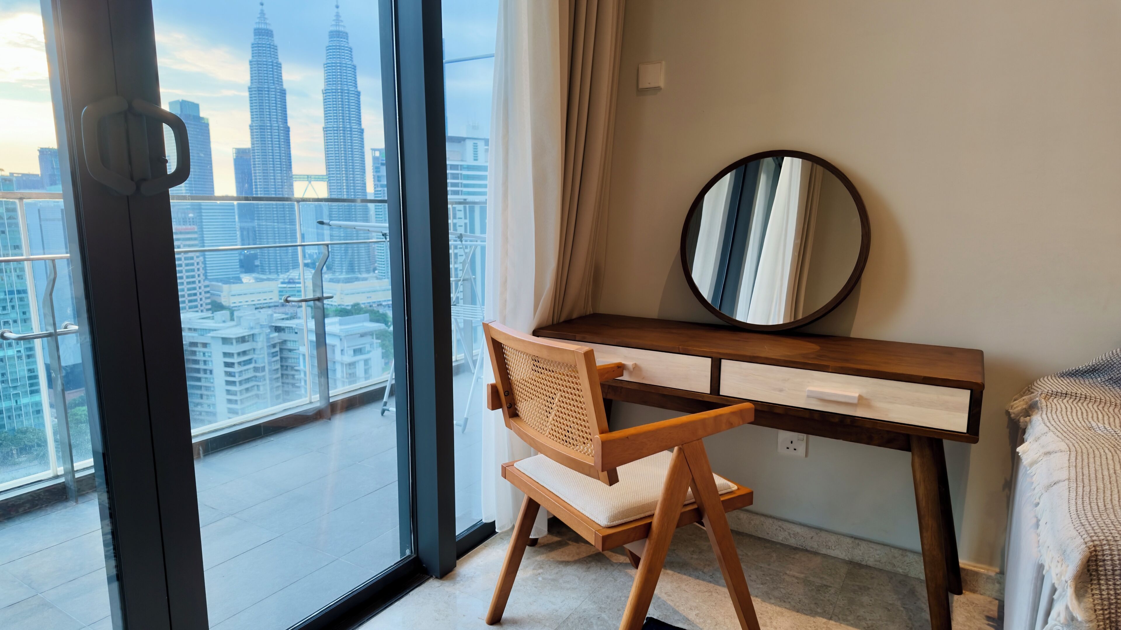 A modern bedroom with a wooden desk, a chair, a round mirror, and a view of a cityscape through large windows.