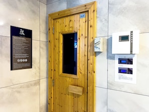 Porta de sauna com painel de madeira e janela de vidro, um aviso com orientações para a sauna e um painel de controle com visor digital.