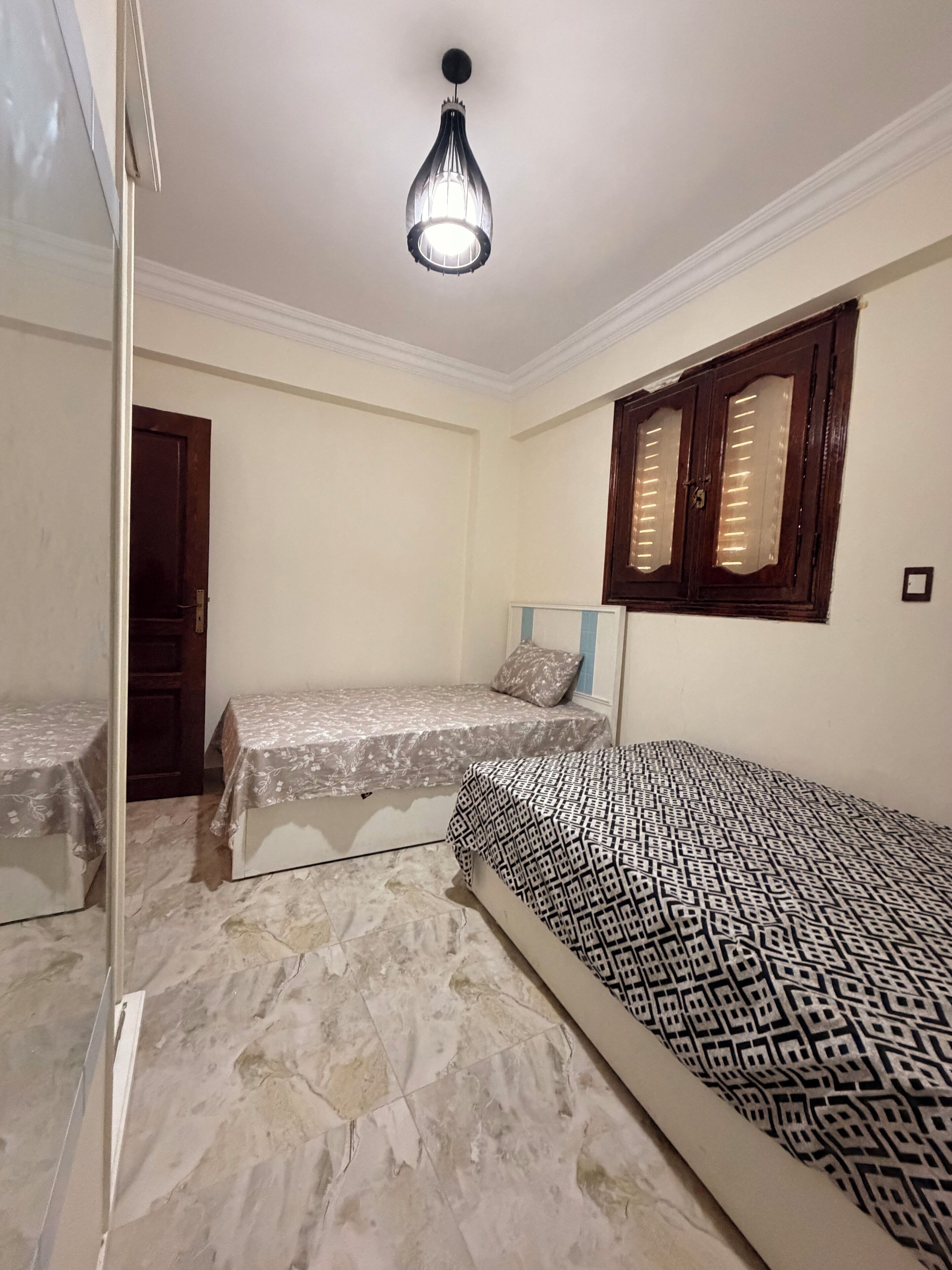 Une chambre avec deux lits simples, une armoire, une fenêtre avec volets et un luminaire suspendu.