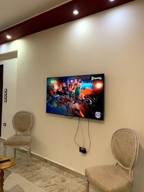 A television mounted on a wall displaying a colorful scene, with two chairs and a table in the foreground. - Chalet in Horus Village and Golden Sands, North Coast (North Coast)