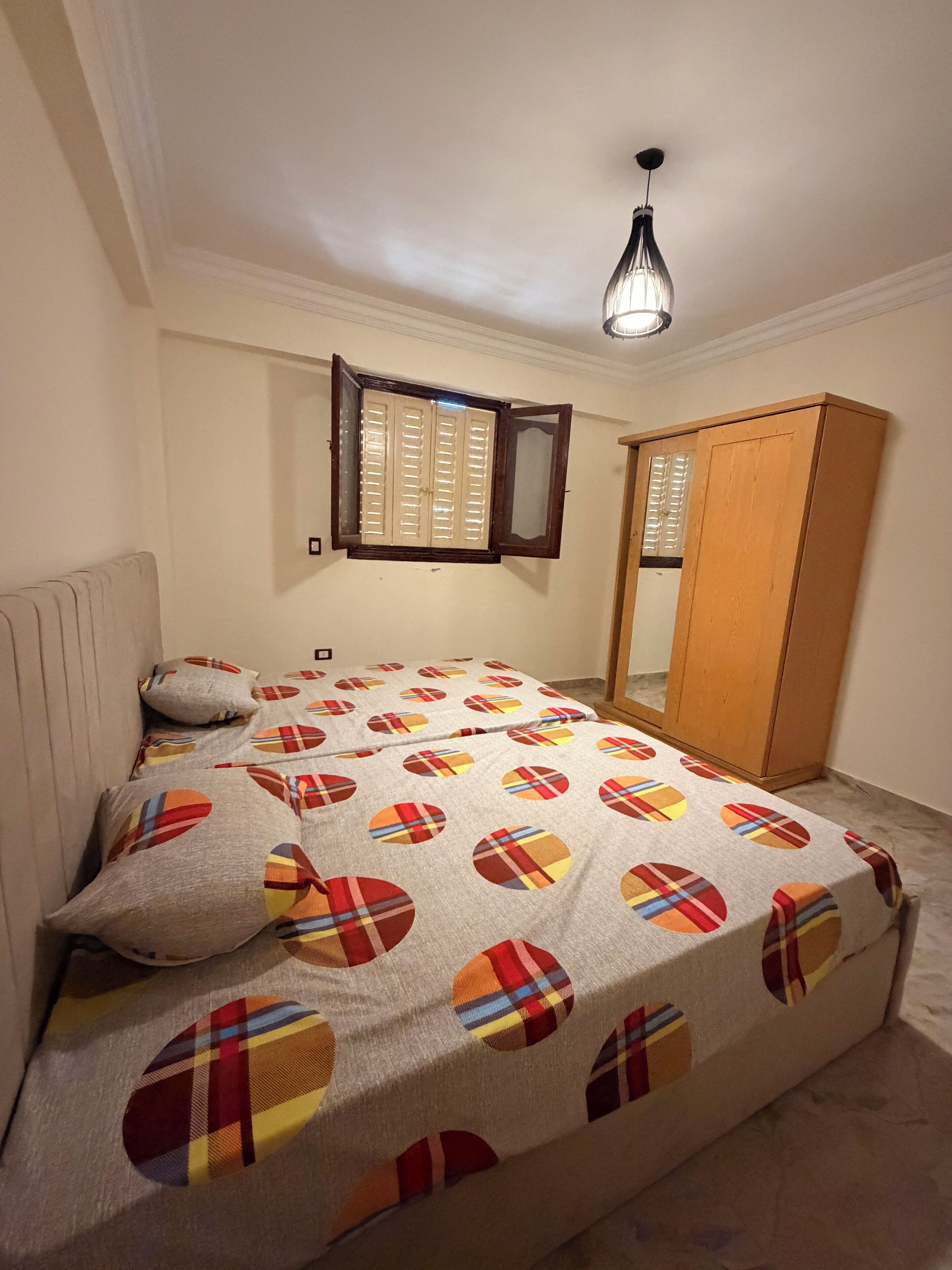 Un dormitorio con cama, ventana con contraventanas, armario y lámpara colgante.