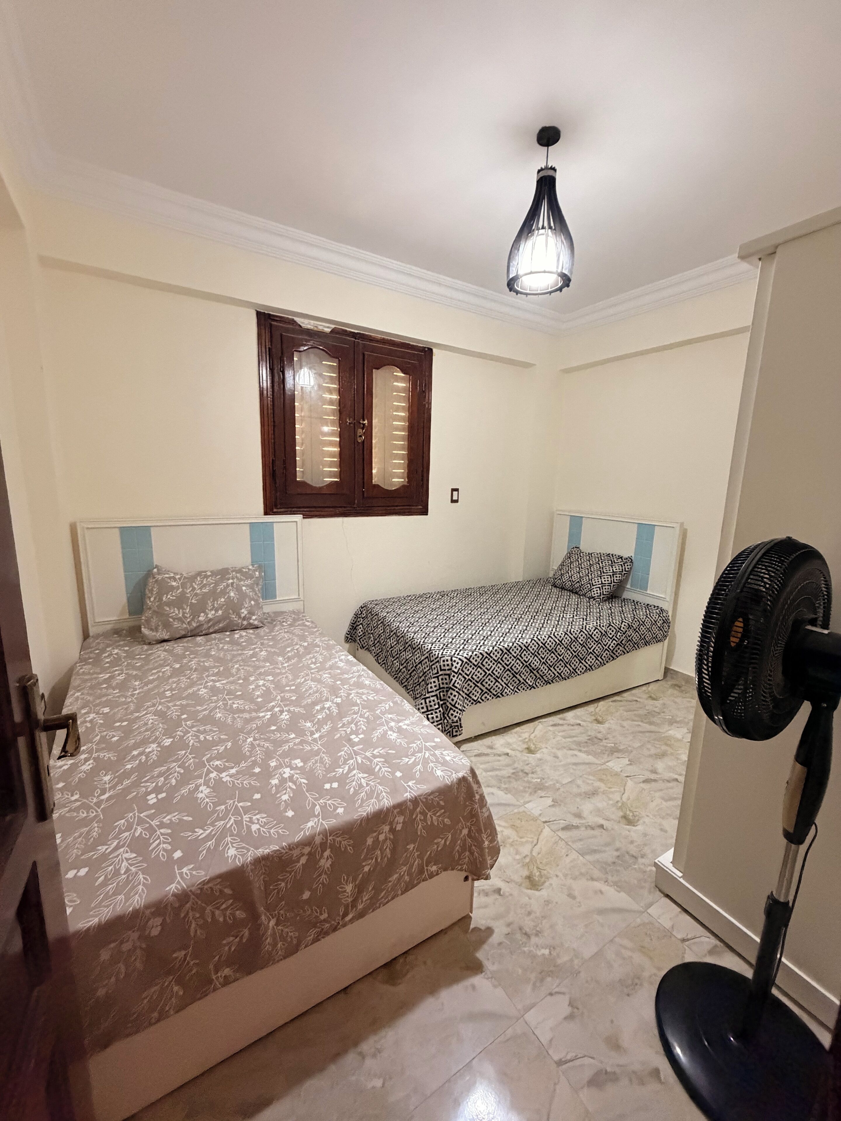 A bedroom with two beds, a ceiling fan, and a window with wooden shutters.