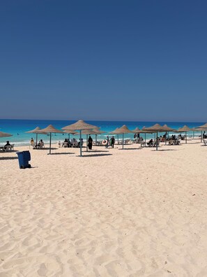 A beach with thatched umbrellas and sun loungers, a clear blue sky, and the ocean in the background.