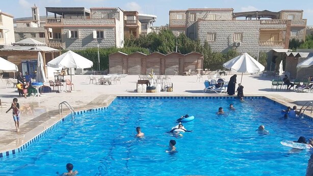 Une piscine avec des personnes dans l’eau, entourée de bâtiments et de coins salons extérieurs.