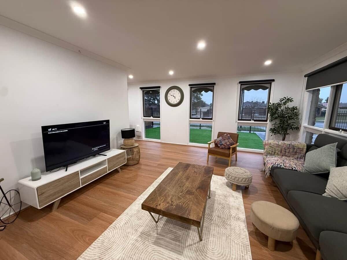 A modern living room with a flat-screen TV, wooden coffee table, sofa, and a large window offering a view of a green lawn.