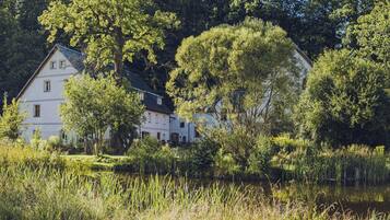 A white building with multiple windows, surrounded by lush greenery and a body of water.