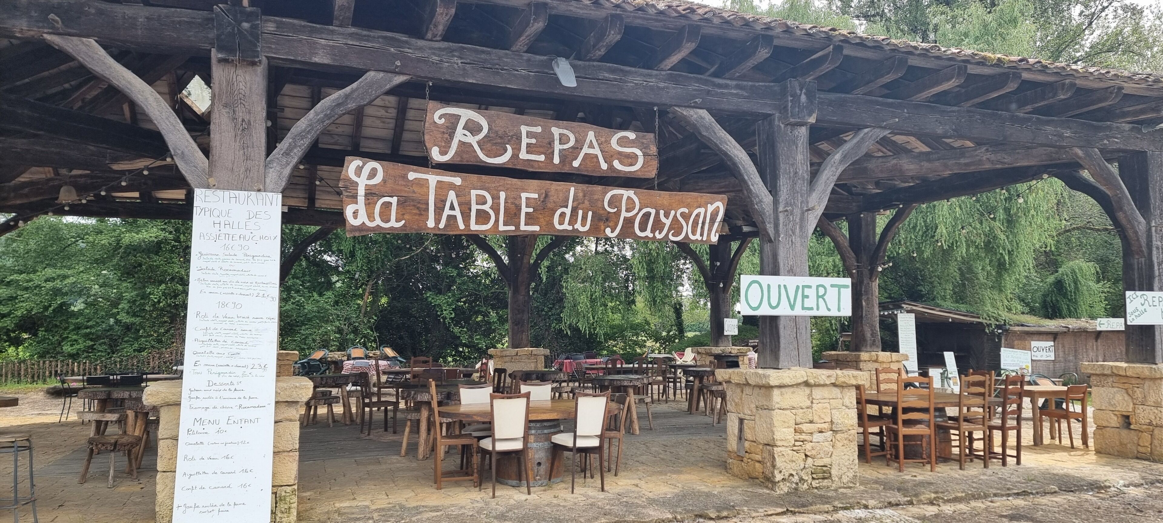 Un restaurante al aire libre con mesas y sillas de madera, un panel con la carta y un letrero que indica que está abierto.