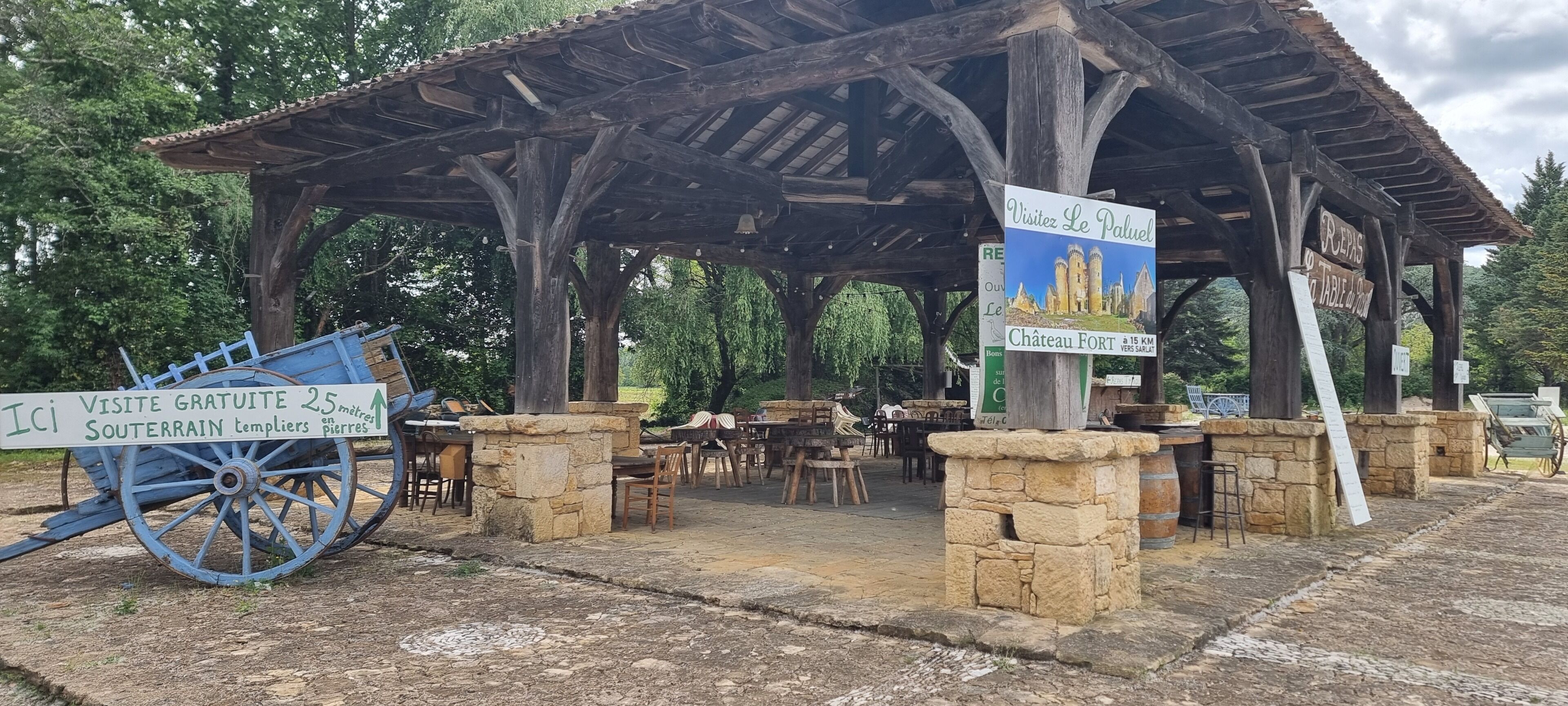 Un pabellón rústico de madera con pilares de piedra, un cartel que anuncia una atracción cercana y asientos al aire libre.
