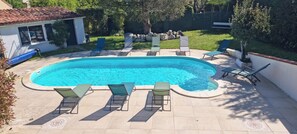 Ein rechteckiger Swimmingpool mit einer gefliesten Liegefläche, umgeben von Liegestühlen und einem kleinen Gebäude mit einem Ziegeldach.
