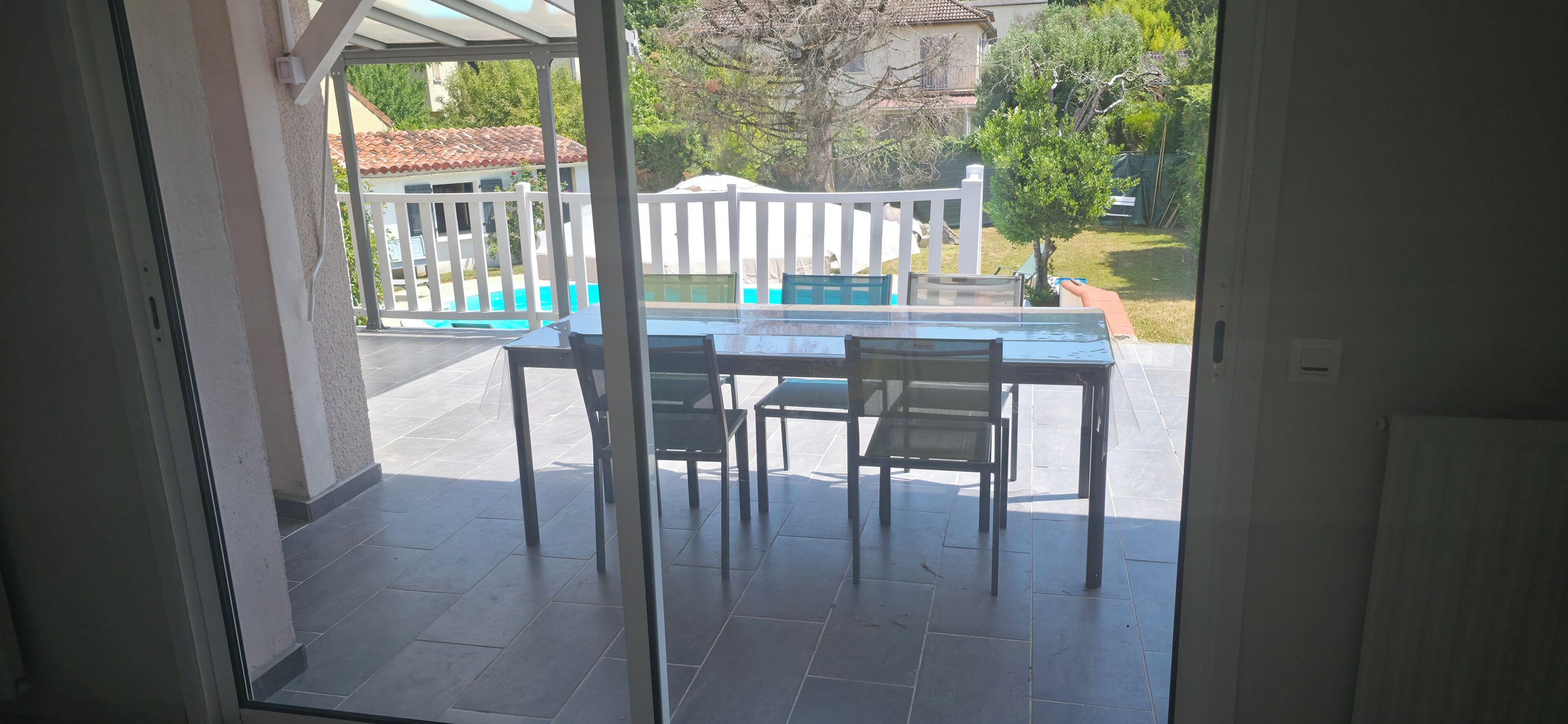Eine Terrasse mit Tisch und Stühlen, ein weißer Zaun sowie Blick auf einen Pool und Grünflächen.