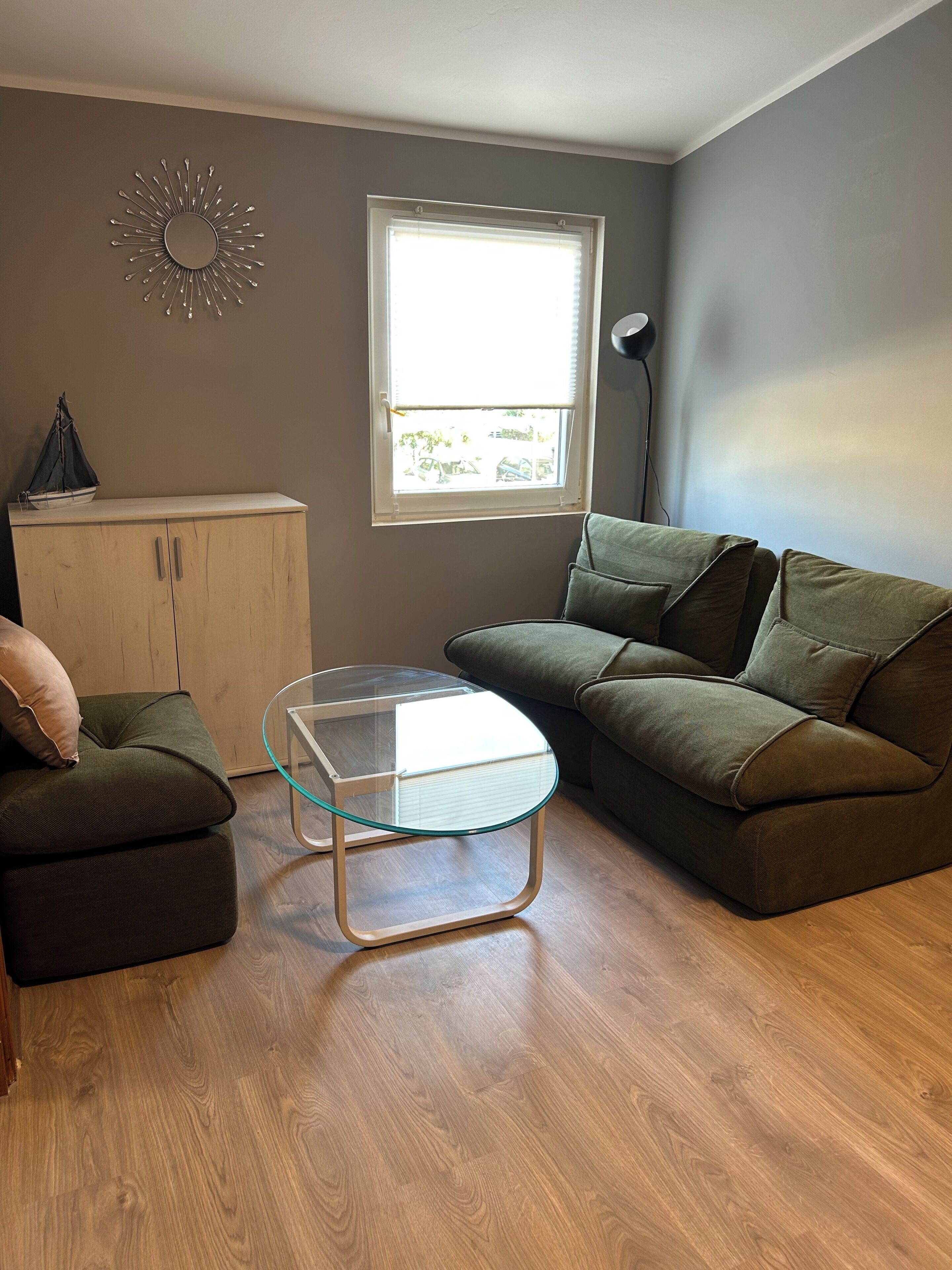 Uma sala de estar com uma mesa de centro de vidro, um sofá verde, um aparador de madeira e uma janela com persianas.