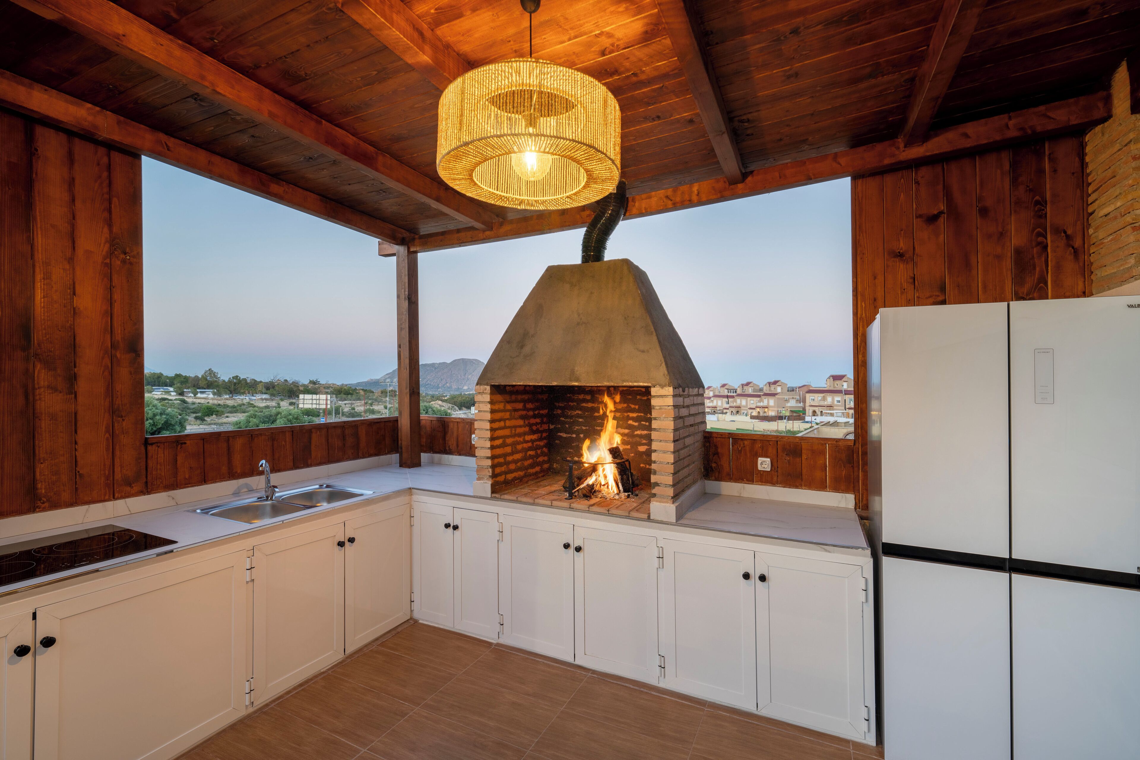 A kitchen with a brick oven, wooden walls, a view of the landscape, and a modern refrigerator.