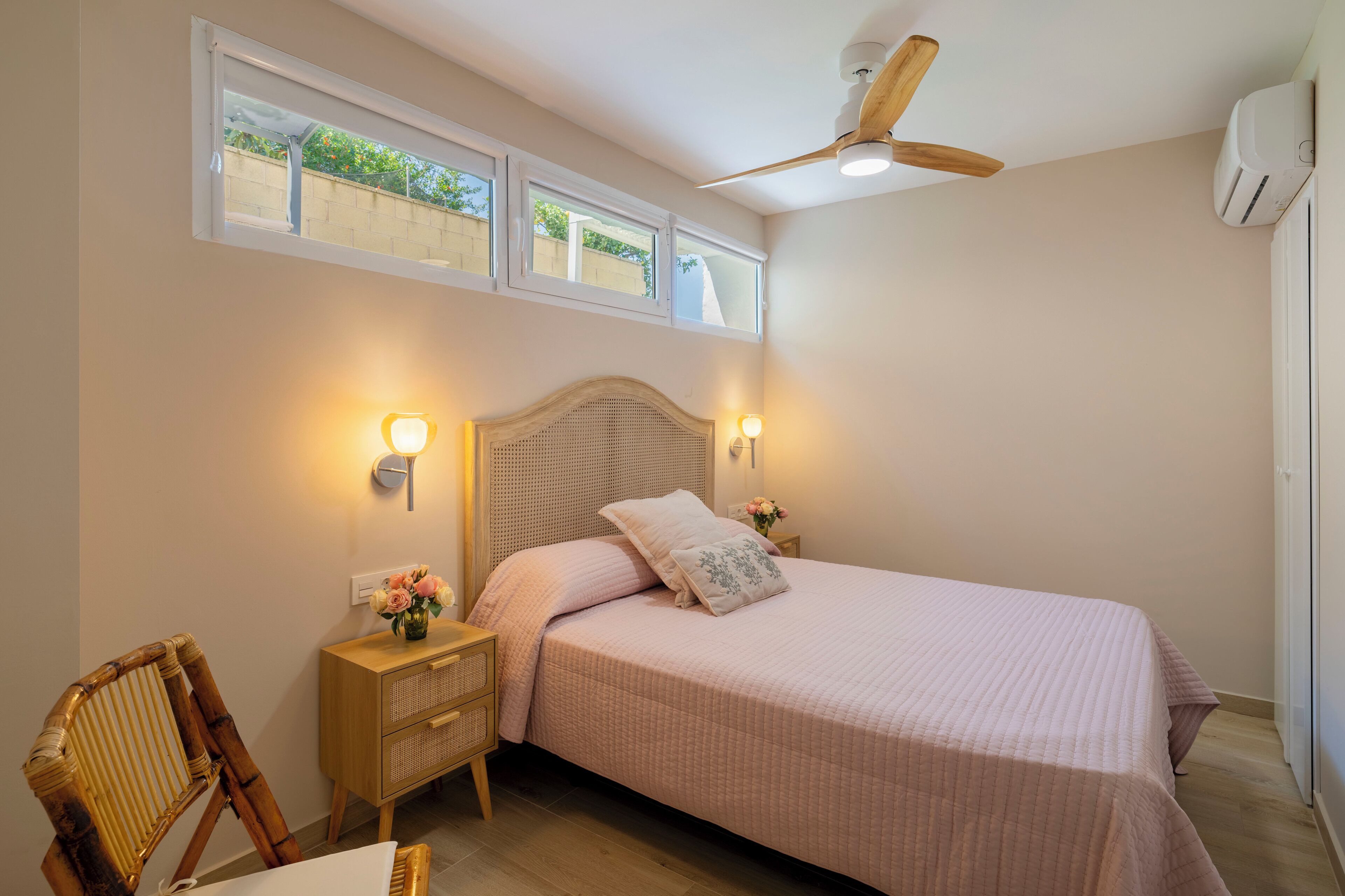 Un dormitorio con cama, lámparas de noche, mesita de noche, una silla, ventilador de techo y ventana.