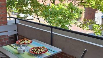 A balcony with a table and chairs, a plate of food, and a view of trees and buildings.