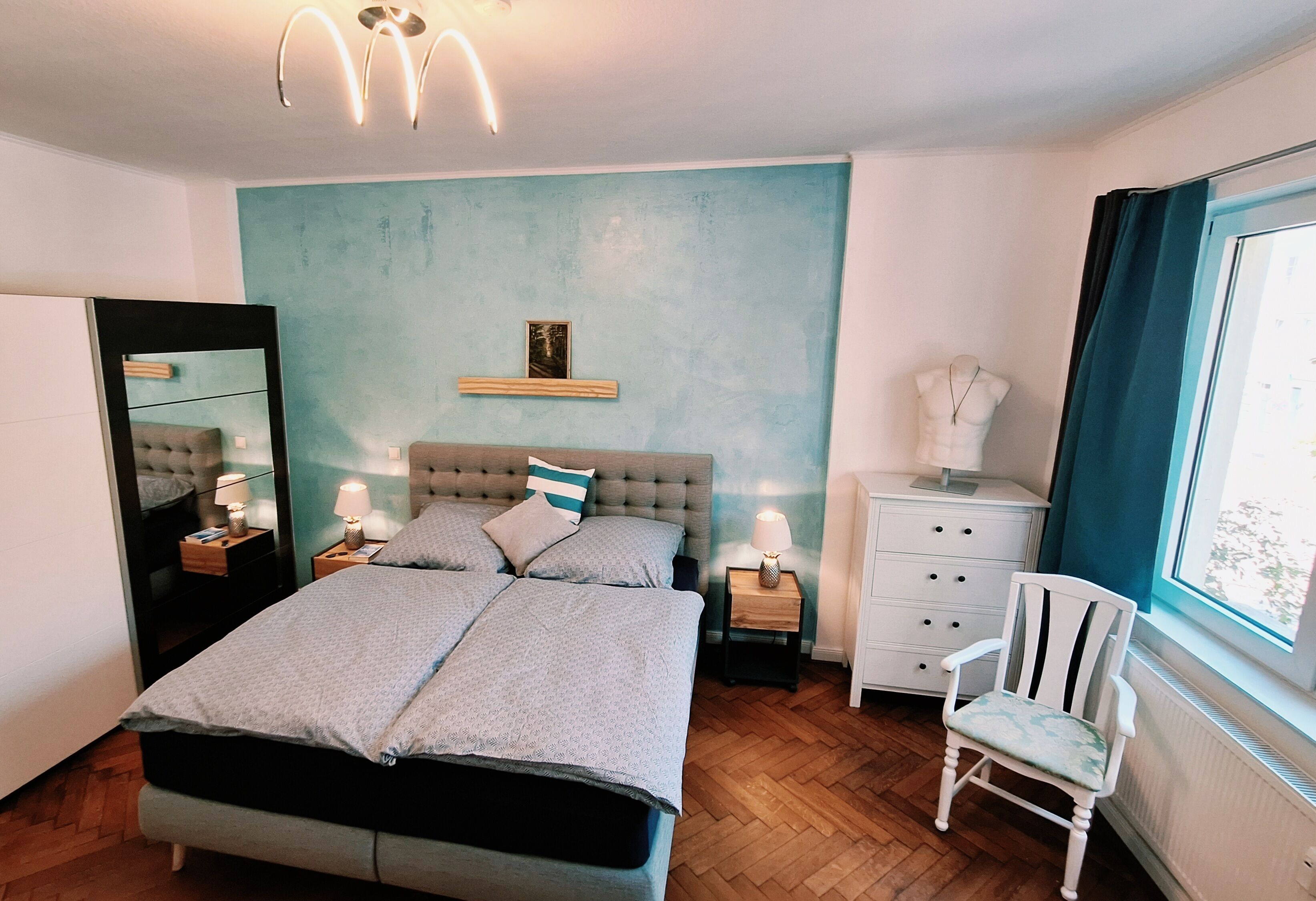 Ein Schlafzimmer mit Doppelbett, Nachttischen mit Lampen, einem Schminktisch, einem Sessel, einem Spiegel und einem Bild an der Wand.