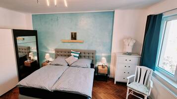Ein Schlafzimmer mit Doppelbett, Nachttischen mit Lampen, einem Schminktisch, einem Sessel, einem Spiegel und einem Bild an der Wand.