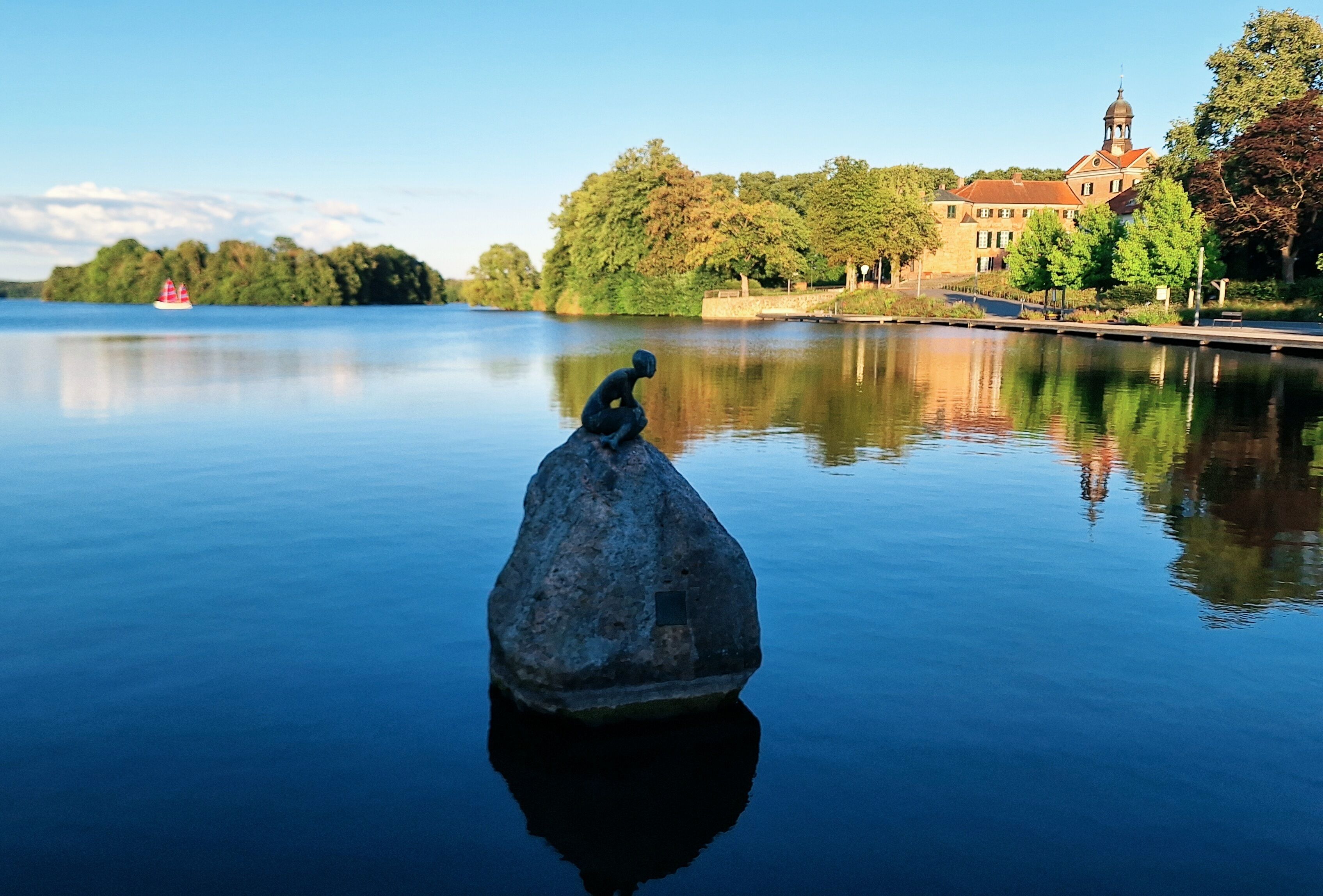 A calm lake with a statue in the foreground and a building with a dome in the background.