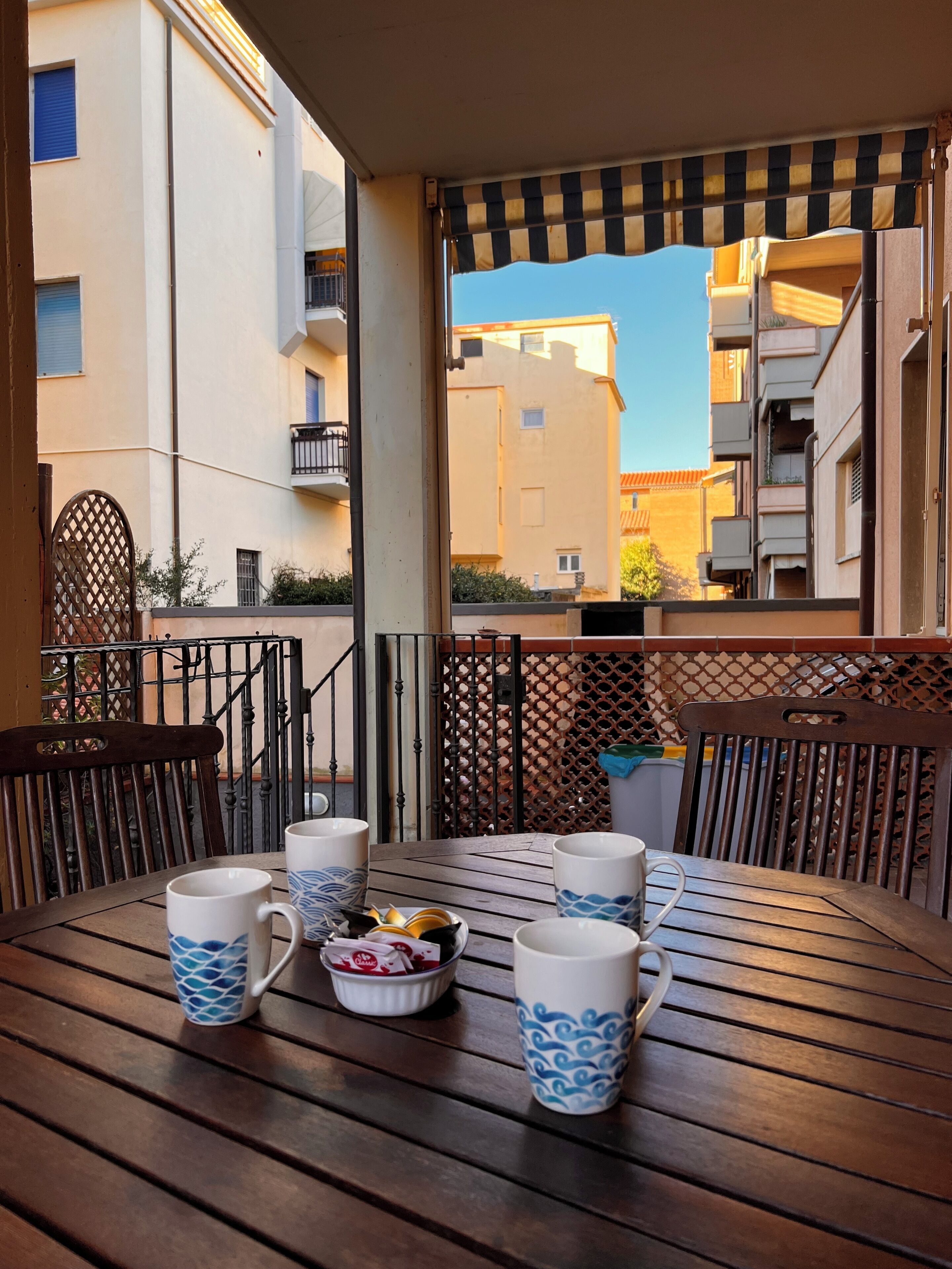 Una mesa de madera con cuatro tazas decoradas y un pequeño plato de aperitivos, frente a una barandilla de balcón con vistas a una zona residencial con edificios y un cielo despejado.