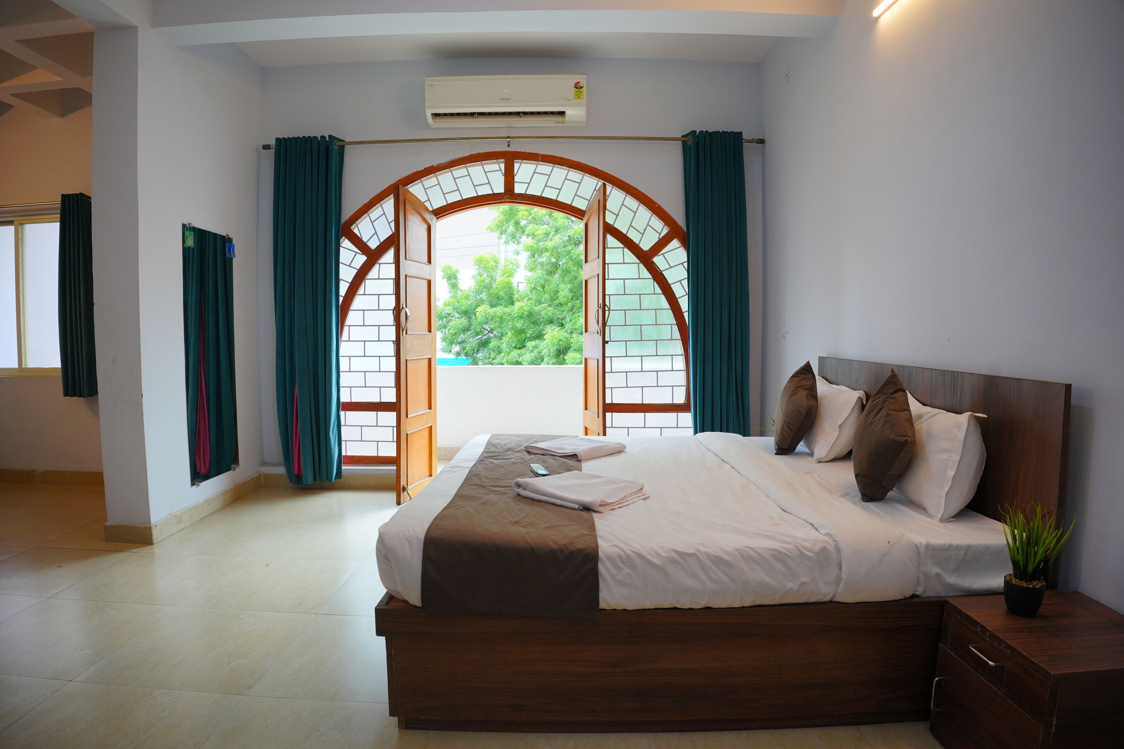 Una camera d'albergo con un letto grande, una testiera in legno, un comodino, una pianta in vaso e vista attraverso una finestra ad arco.